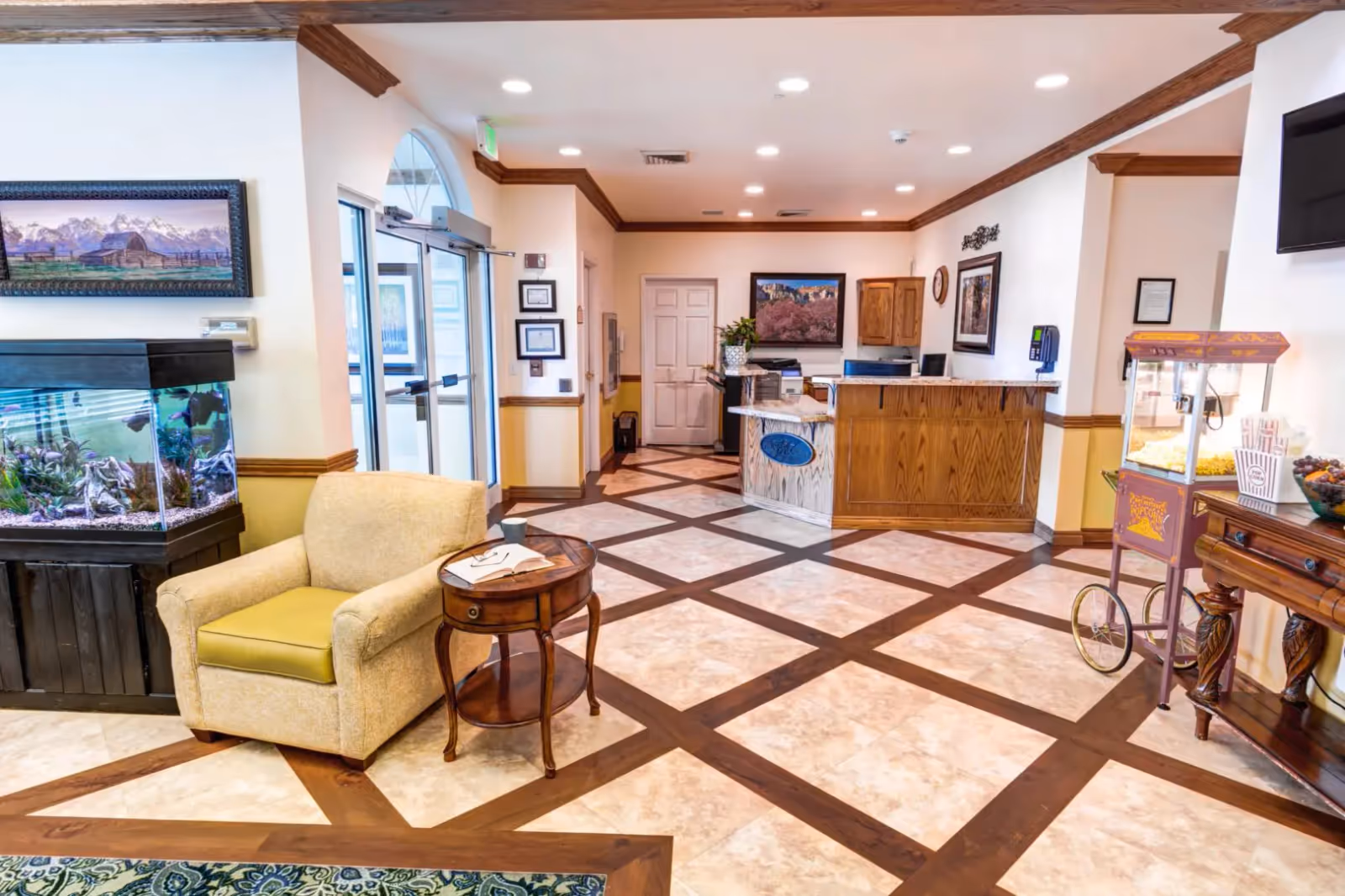 Interior view of a senior living facility lobby with a beige armchair and wooden side table, a large fish tank, a popcorn machine on a wooden table, and a reception desk in the background. The floor has a tiled pattern with wooden accents, and the walls are decorated with framed pictures.
