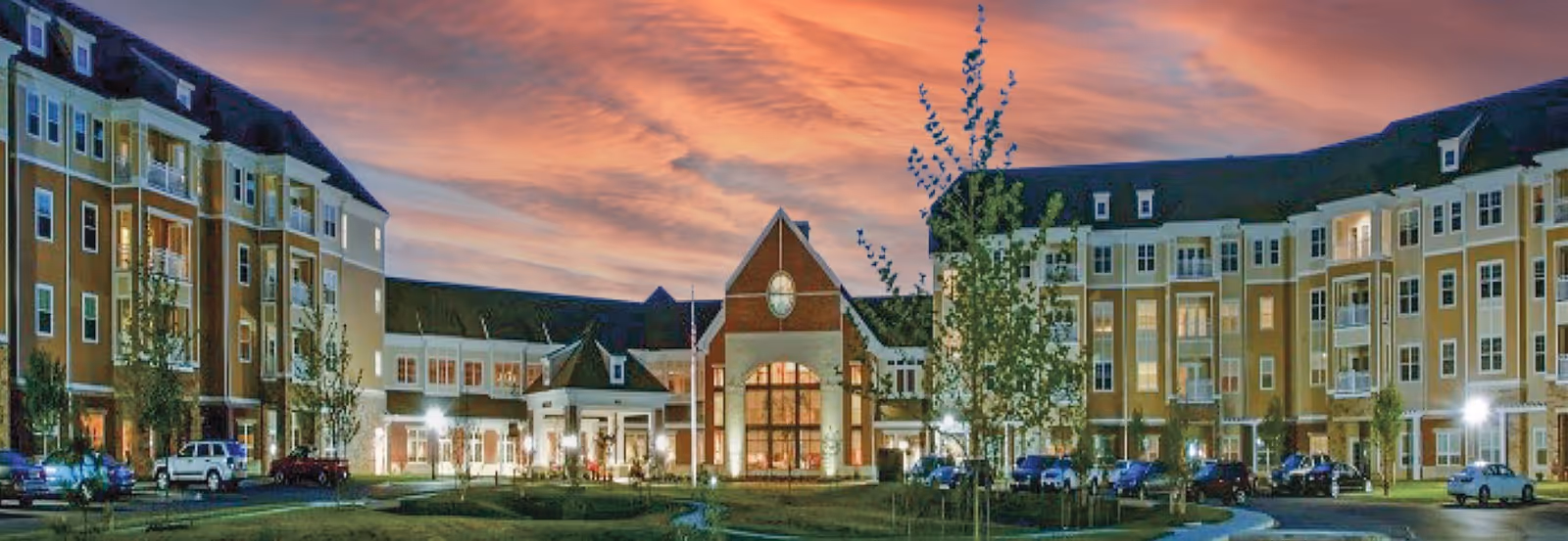 Large multi-story senior living building with a central glass entrance, surrounding wings, parked cars, and a colorful sunset sky.