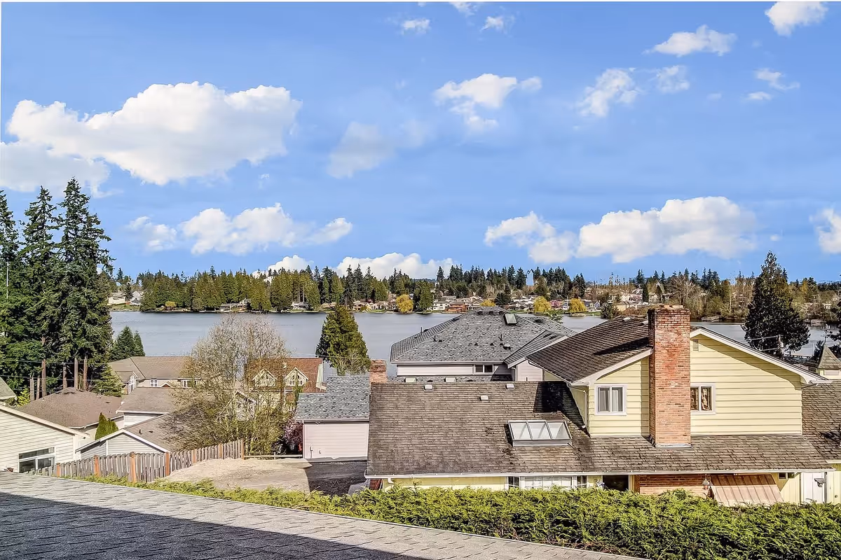 View over rooftops of houses with a lake and trees in the background under a blue sky with scattered clouds.