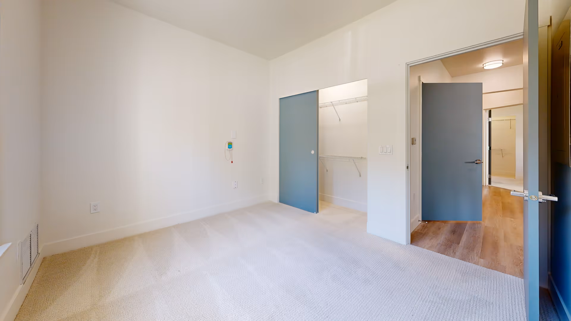 Empty bedroom with beige carpet, white walls, a blue sliding closet door partially open revealing a closet with wire shelving, and a blue door open to a hallway with wood flooring.