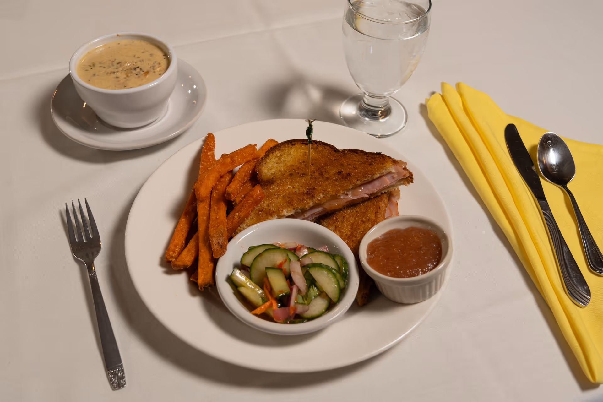 A meal served on a white plate consisting of a grilled ham and cheese sandwich, sweet potato fries, a small bowl of cucumber salad, and a small bowl of applesauce. Next to the plate is a cup of creamy soup on a saucer, a glass of water, and a set of silverware wrapped in a yellow napkin on a white tablecloth.