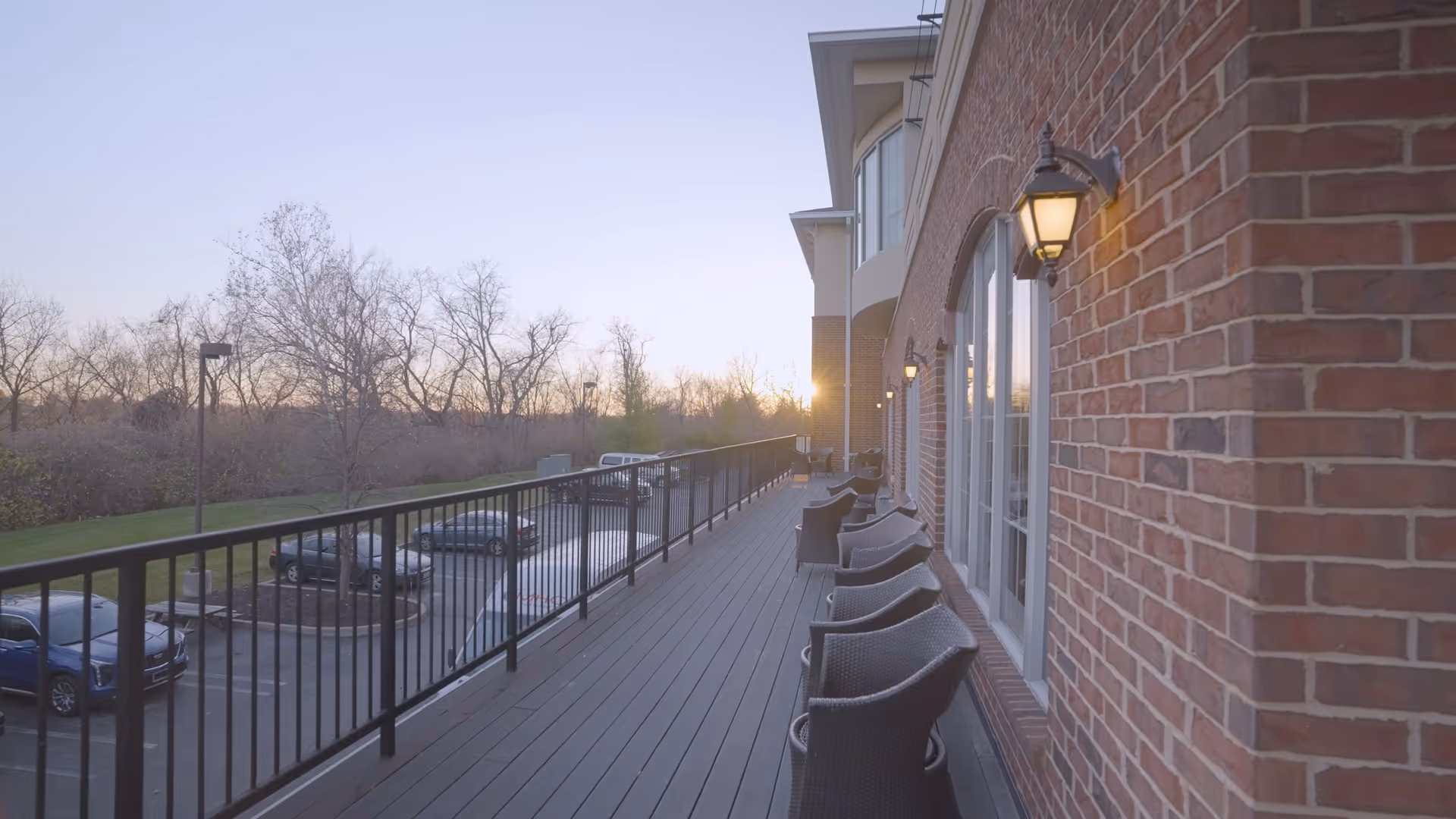 Outdoor balcony area of a brick building with several wicker chairs lined up along the railing, overlooking a parking lot with cars and leafless trees in the background during sunset.