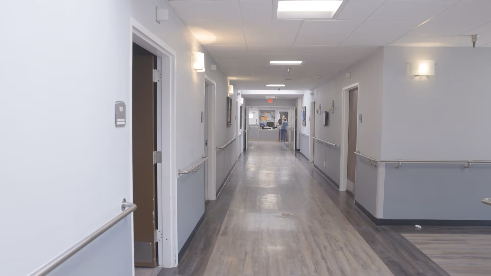 A long, clean hallway in a healthcare facility with light gray walls, handrails on both sides, several doors leading to rooms, and a reception desk at the far end where a person is standing.