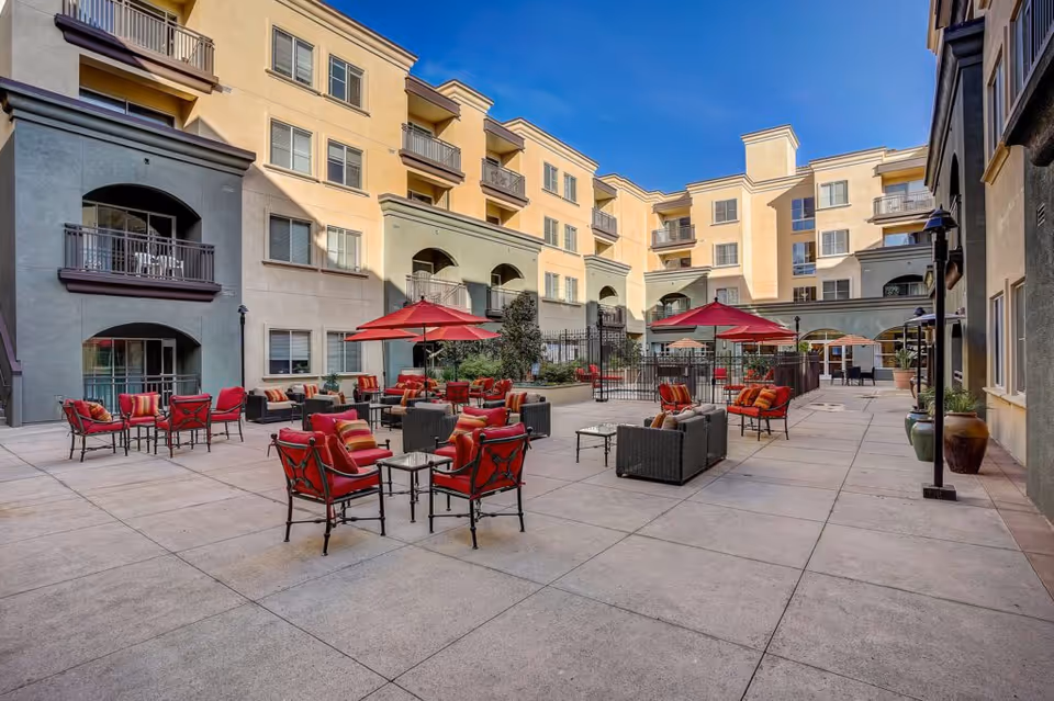 Spacious sunlit courtyard with red umbrella-shaded seating surrounded by a multi-story apartment building with balconies.