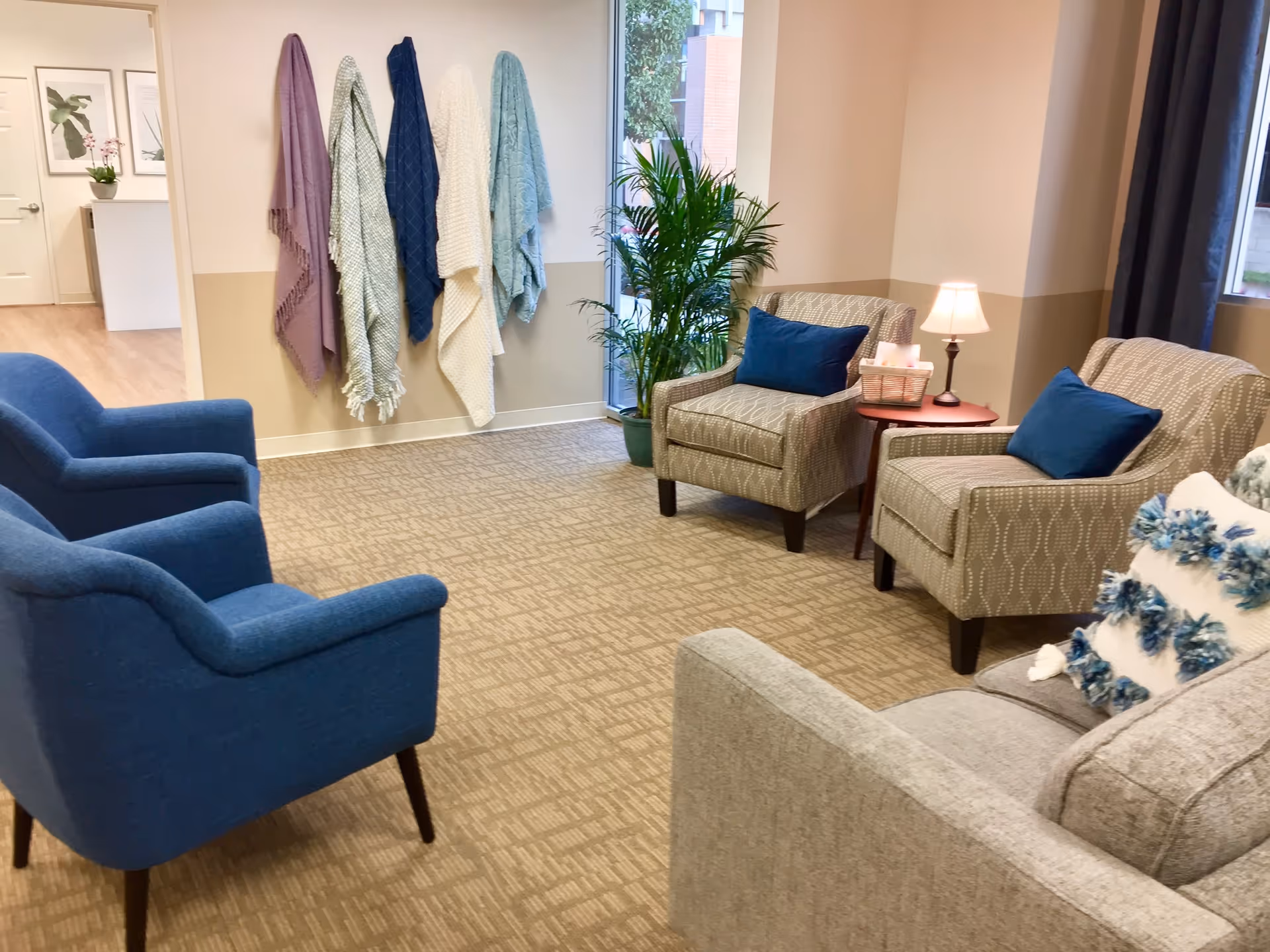 Cozy common sitting room with blue and patterned armchairs, throw blankets hanging on the wall, a potted plant and a side table with a lamp.