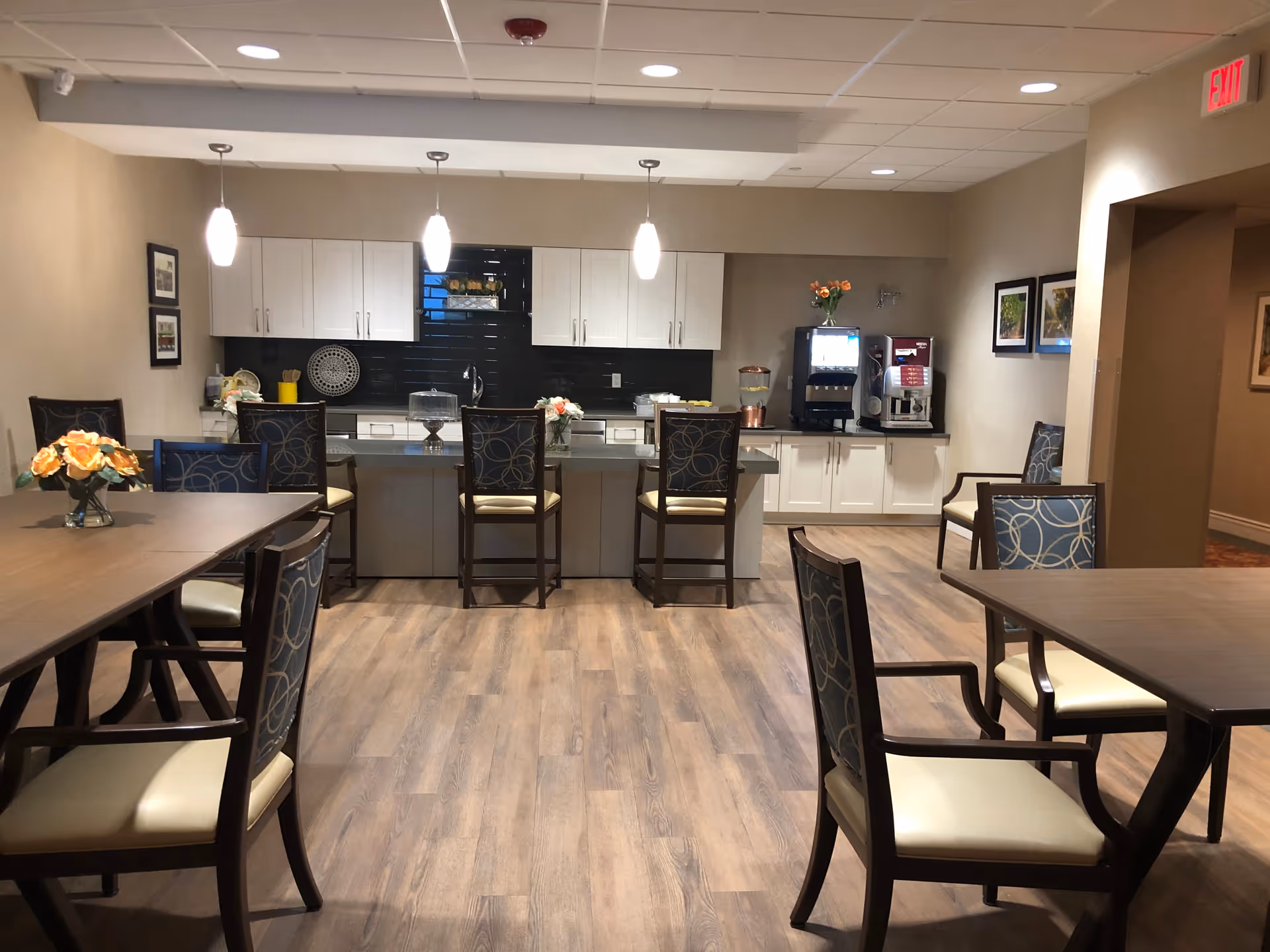 A senior living facility dining area with wooden tables and cushioned chairs arranged neatly. In the background, there is a kitchen counter with bar stools, white cabinets, a black backsplash, and pendant lights hanging from the ceiling. On the counter, there are flowers, a cake stand, and beverage dispensers. The room has wood flooring and framed pictures on the walls.