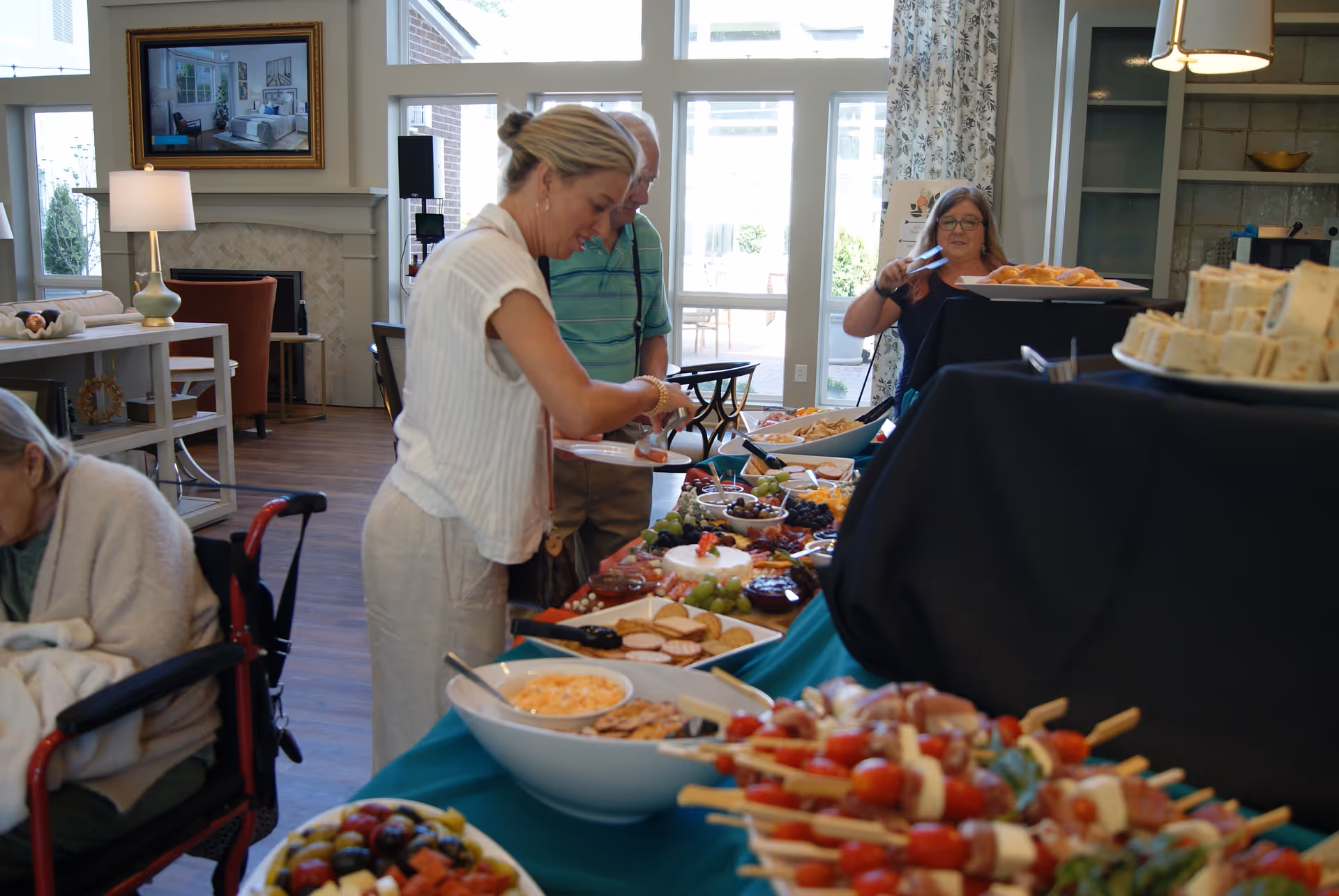 People serving themselves from a buffet of appetizers set up in a bright common room with seating and large windows.