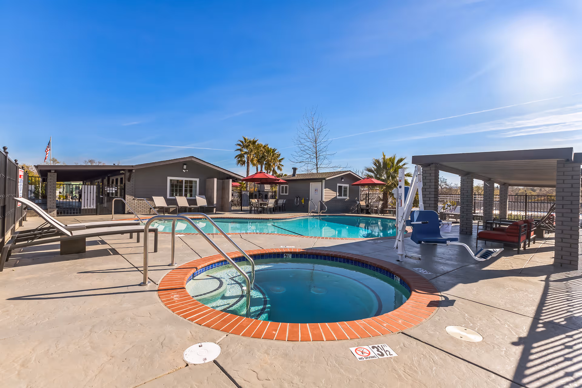 Outdoor community pool and hot tub area with lounge chairs, shaded seating, and surrounding clubhouse buildings under a clear blue sky.