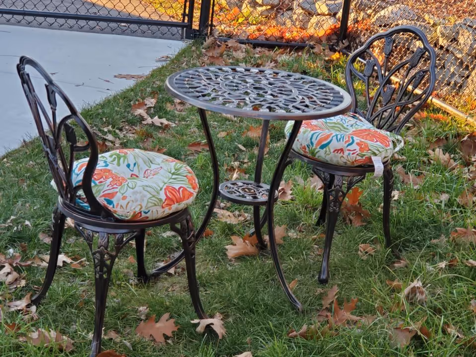 A small outdoor metal table with intricate designs and two matching metal chairs with colorful floral cushions, placed on grass with fallen autumn leaves near a chain-link fence.
