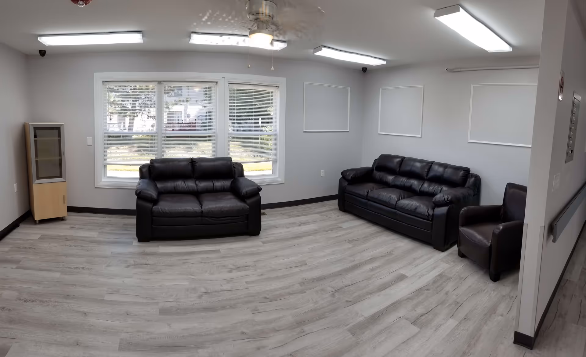 Bright common lounge with two black leather sofas, a leather armchair, a window, and light wood flooring.