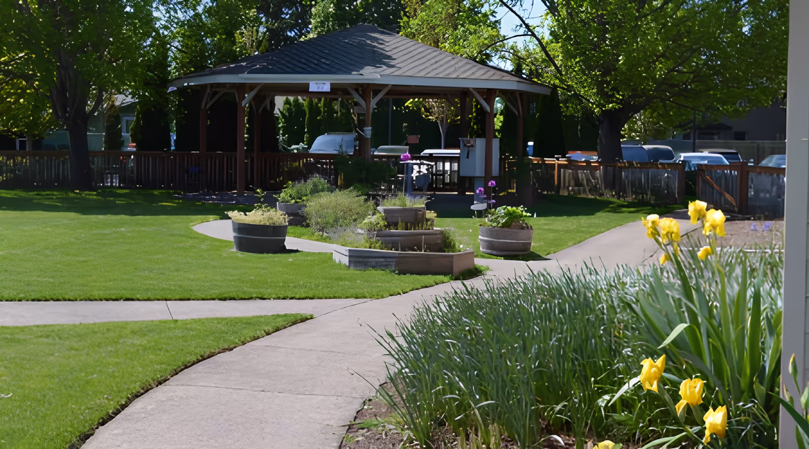 A well-maintained outdoor garden area with a paved walkway leading to a wooden gazebo surrounded by green grass, flower beds, and trees. Yellow flowers and other plants are visible along the path.