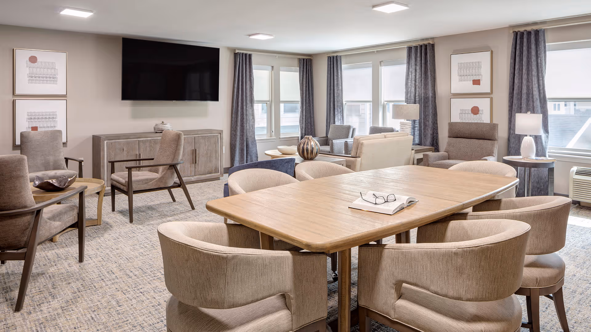 A bright and spacious living room area with a large wooden table surrounded by eight beige upholstered chairs. The room features multiple seating areas with armchairs and sofas, a wall-mounted flat-screen TV above a wooden cabinet, large windows with gray curtains, and modern lamps providing ambient lighting.