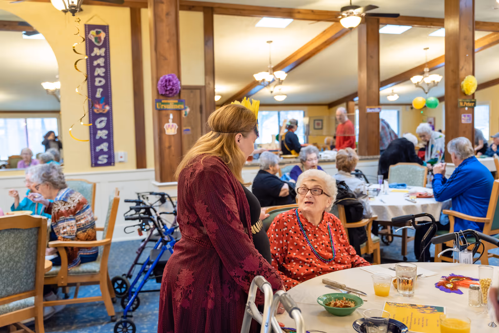 A lively dining room in a senior living facility decorated for Mardi Gras, with elderly residents seated at round tables enjoying meals and socializing. A woman wearing a red dress and a yellow crown is standing and talking to an elderly woman in a red polka dot blouse seated at a table. The room has wooden beams, ceiling fans, and festive decorations including balloons and Mardi Gras banners.