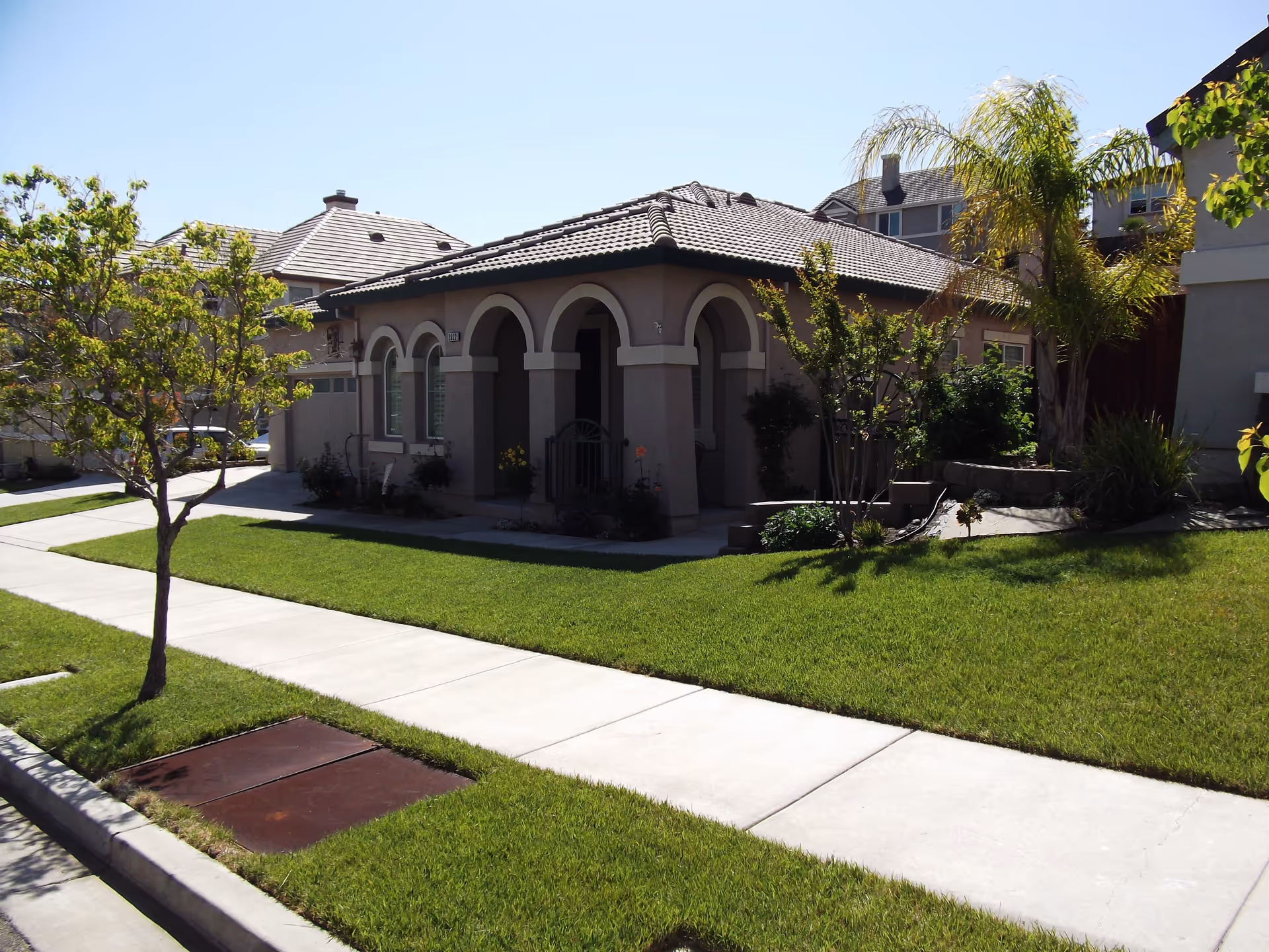 Front view of a single-story stucco house with an arched porch, manicured lawn, sidewalk, and small trees.