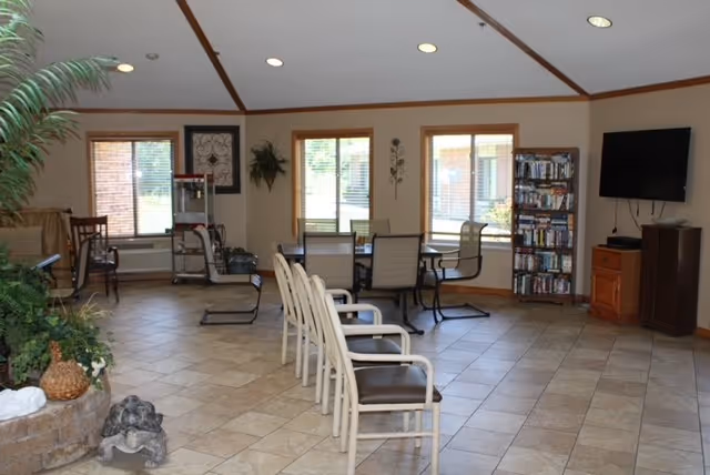 Spacious senior living common room with a row of chairs, tables, a wall-mounted TV, bookshelf and large windows.