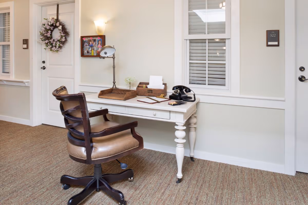 A small white desk with a vintage black rotary phone, a desk lamp, and wooden organizers sits against a light-colored wall. A brown cushioned office chair with wooden armrests is positioned in front of the desk. There are two white doors on either side of the desk, one decorated with a floral wreath and the other labeled with the number 322. A window with white shutters is above the desk, and a framed photo collage hangs on the wall.