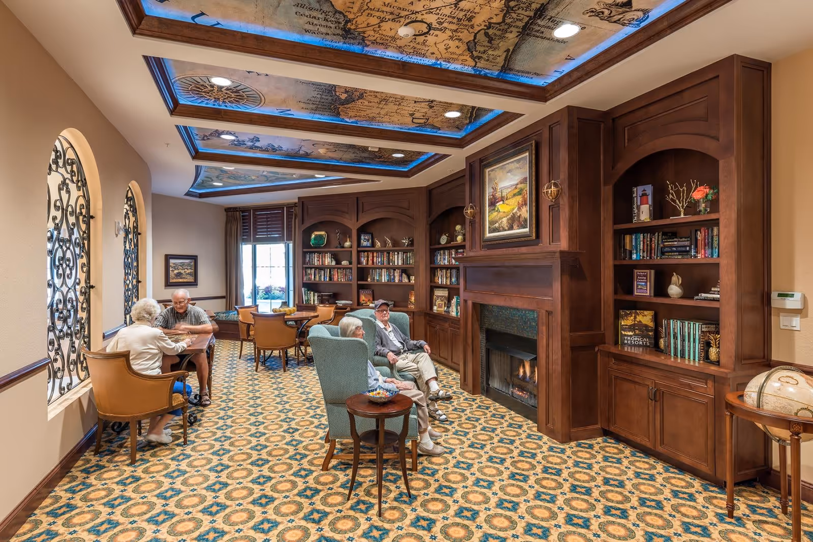 A cozy common area in a senior living facility with patterned carpet, wooden bookshelves filled with books and decorations, a fireplace with a painting above it, and a ceiling decorated with illuminated map panels. Four elderly people are seated, two at a table playing a game and two relaxing in armchairs near the fireplace.