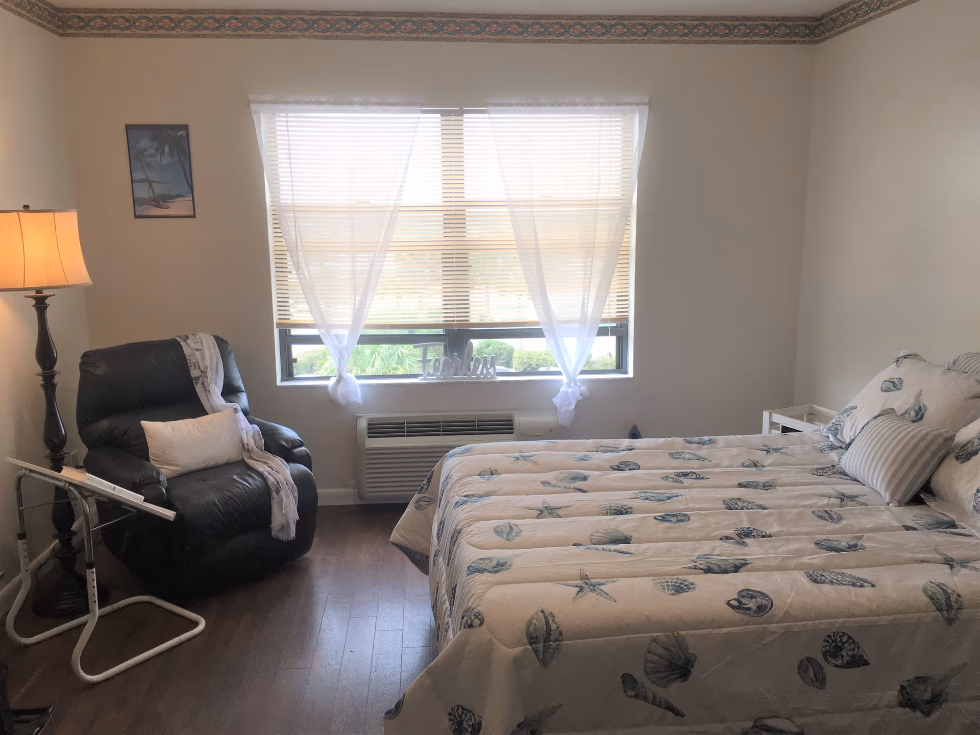 A cozy bedroom with a large bed covered in a seashell-patterned comforter and matching pillows. Next to the bed is a small white nightstand. A black recliner chair with a white pillow and a light throw blanket is positioned near a tall floor lamp. A window with white sheer curtains and blinds lets in natural light, with a decorative 'Together' sign on the windowsill. The room has wooden flooring and light-colored walls with a decorative border near the ceiling.