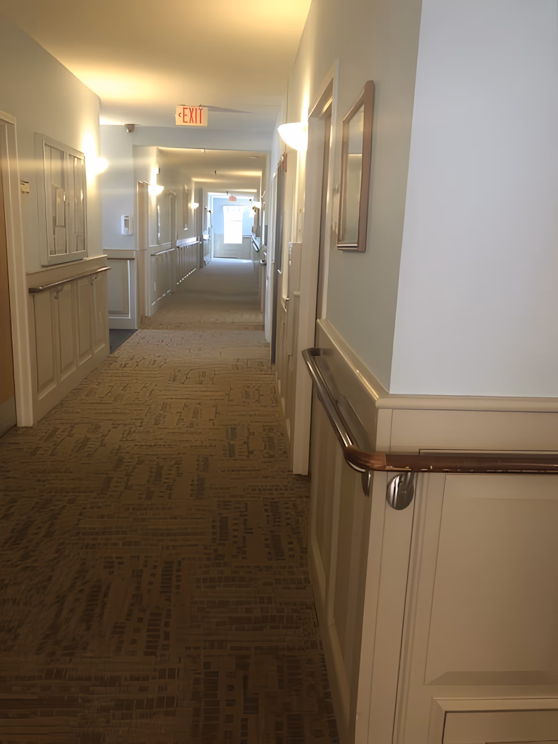 A long, well-lit hallway in a senior living facility with beige patterned carpet, light blue walls, wooden handrails on both sides, and several doors and framed pictures along the walls. An exit sign is visible at the far end of the corridor.