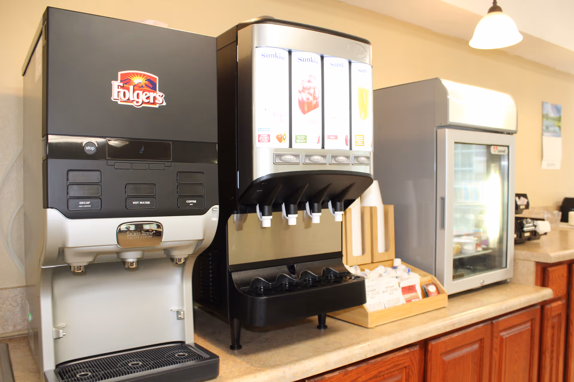 A beverage station with a Folgers coffee machine, a Sunkist juice dispenser with multiple flavors, a small refrigerator, and a countertop with various condiments and napkins in a senior living facility.