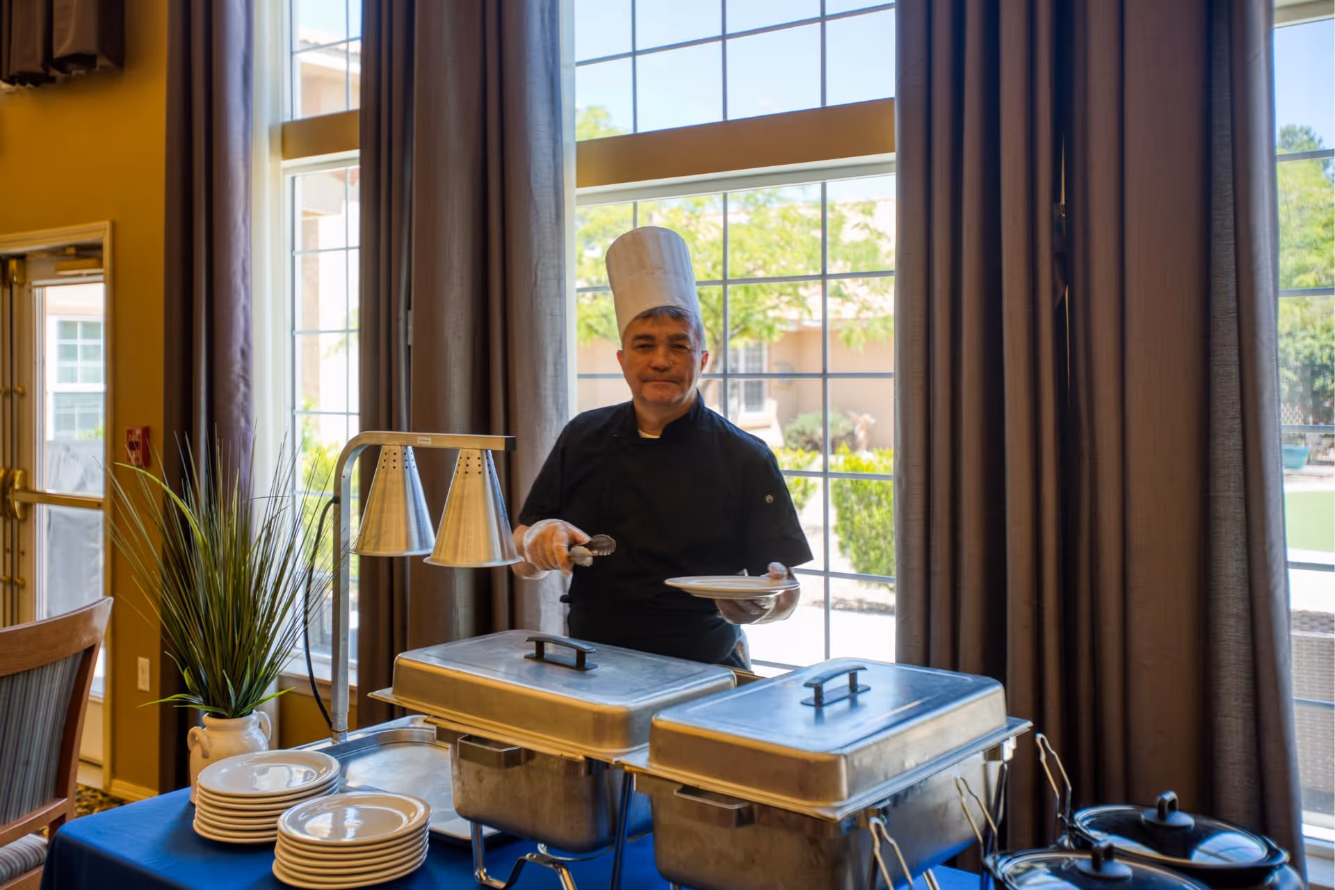A chef wearing a white hat and black uniform stands behind a buffet table with covered chafing dishes and stacks of plates, holding a plate and serving tongs. The setting is indoors with large windows and curtains, and a view of greenery outside.