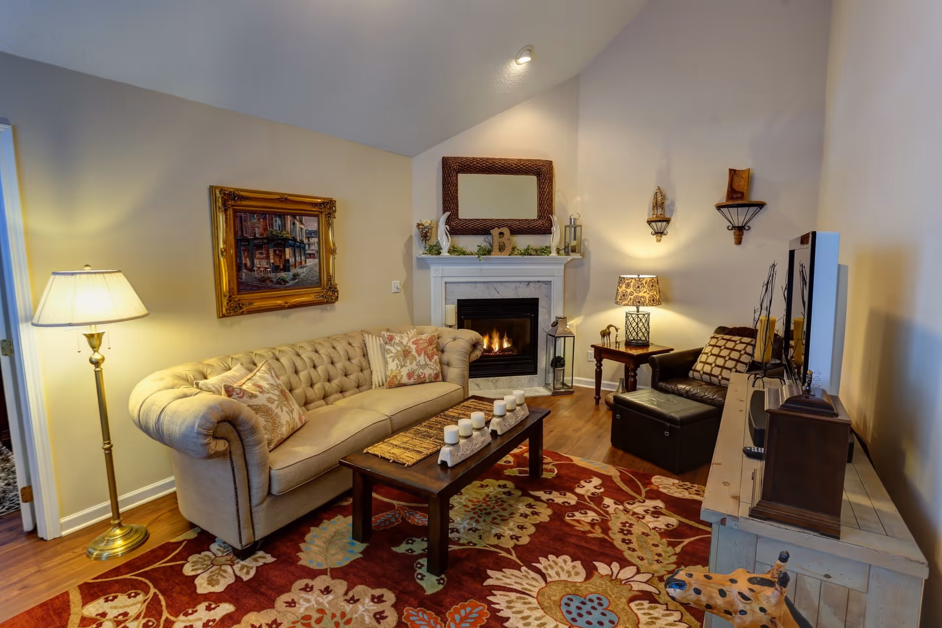 A cozy living room with a beige tufted sofa adorned with floral pillows, a wooden coffee table with white candles, a fireplace with a decorative mirror and ornaments above it, a patterned area rug, a floor lamp, a side table with a lamp and decorative items, a dark leather armchair with a matching ottoman, and a TV on a wooden console.