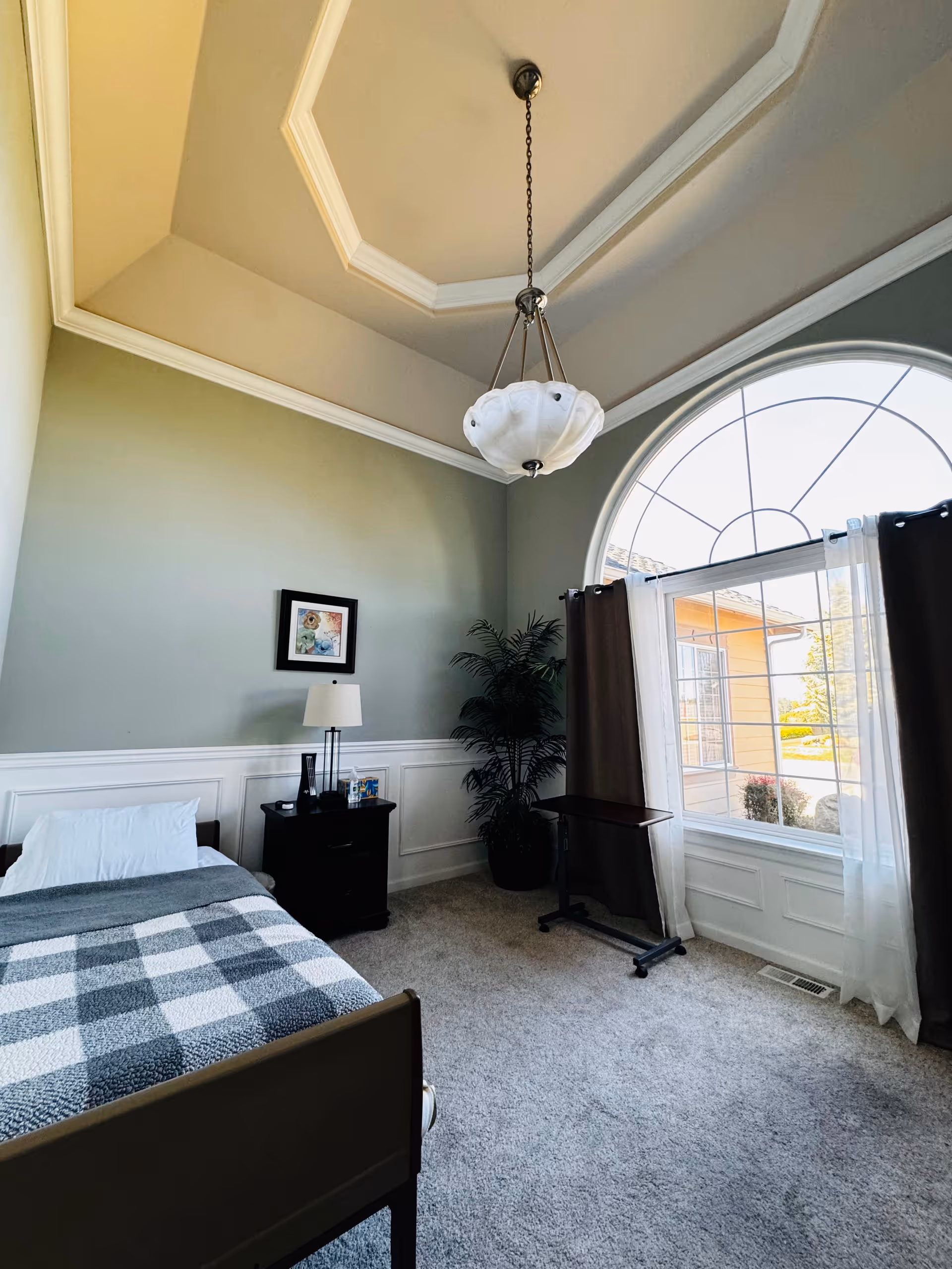 Bright bedroom with a single bed, nightstand and lamp, large arched window with curtains, chandelier, and a potted plant.