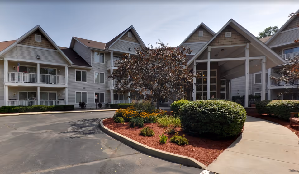 Exterior view of Charter Senior Living of Kenosha showing a multi-story building with balconies, a covered entrance, landscaped bushes, flowers, and a tree in front of the building under a clear sky.