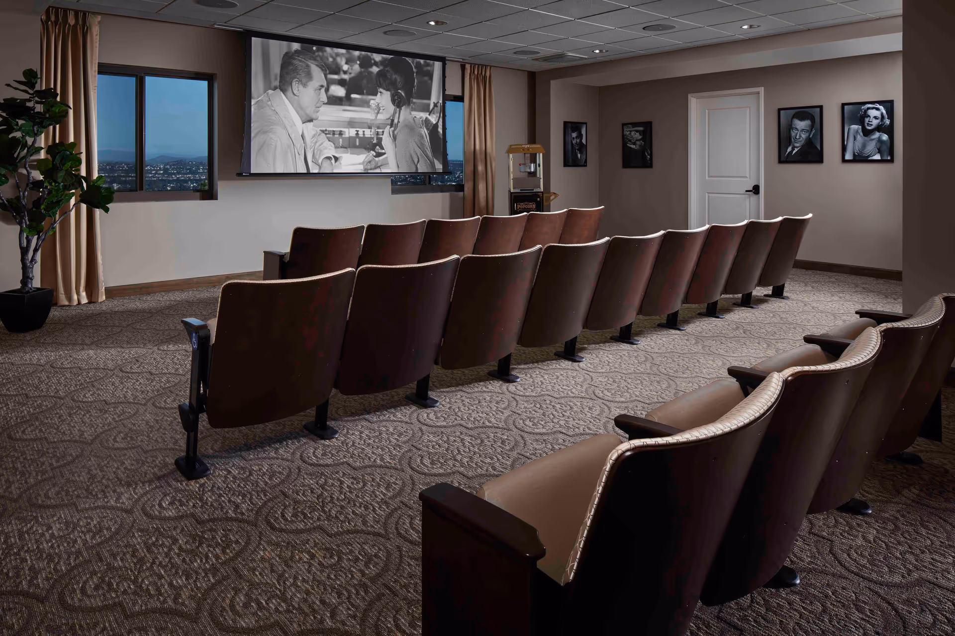 A small theater room with two rows of brown cushioned seats facing a large screen showing a black and white movie scene of a man and woman talking. The room has patterned carpet, beige walls with framed black and white celebrity photos, a popcorn machine in the corner, a window with a city view, and beige curtains.