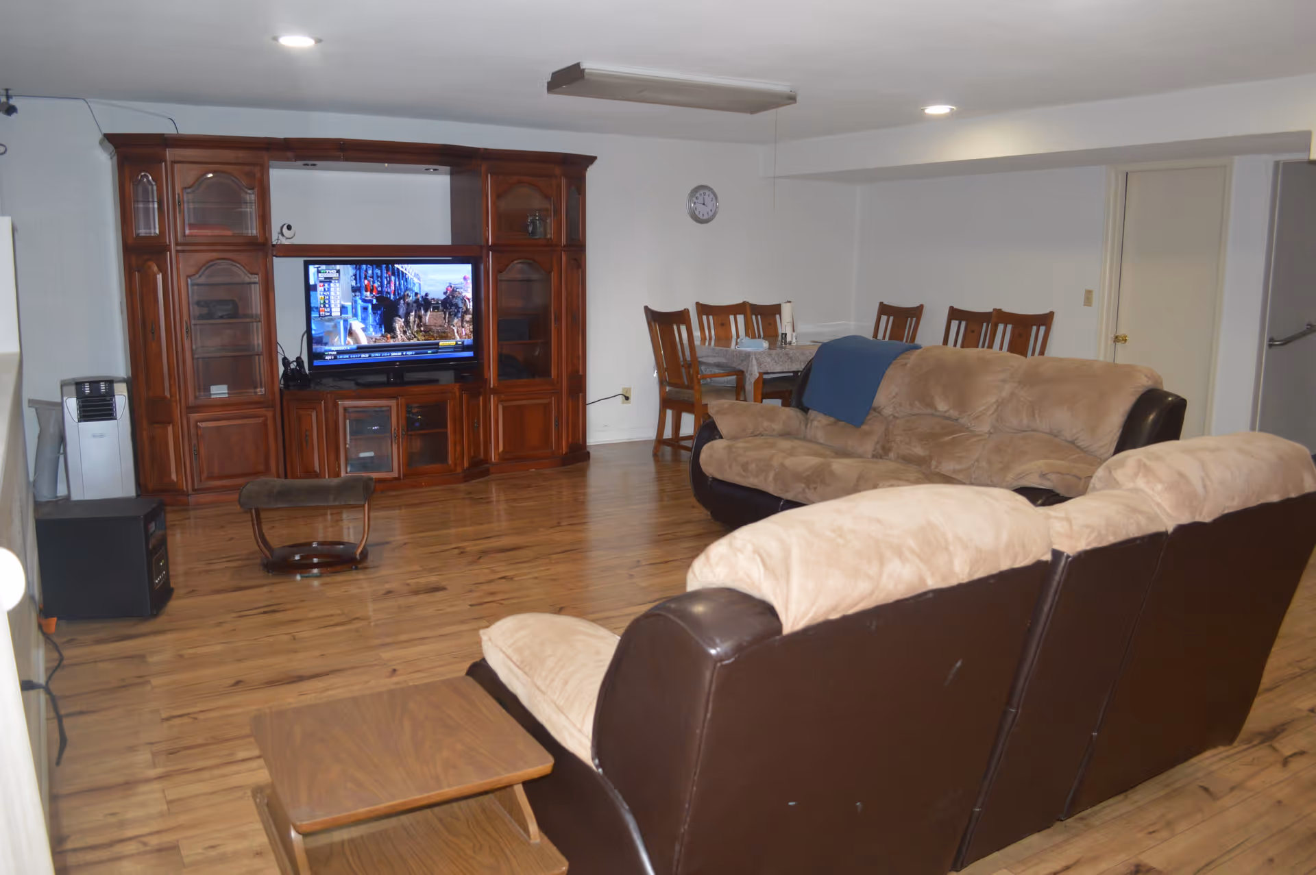 A cozy living room with two beige and brown sofas facing a wooden entertainment center with a flat-screen TV. There is a small wooden coffee table in front of the sofas and a dining table with six chairs in the background. The room has wooden flooring and white walls with a clock on the wall.
