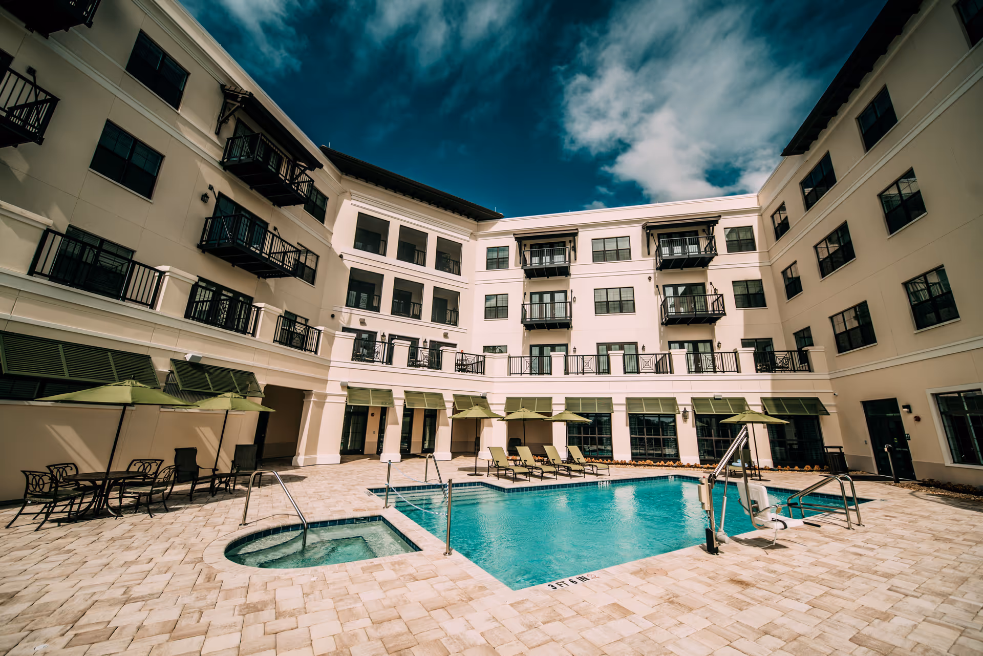 Sunny courtyard featuring a swimming pool and hot tub surrounded by a four-story residential building with balconies and poolside loungers.