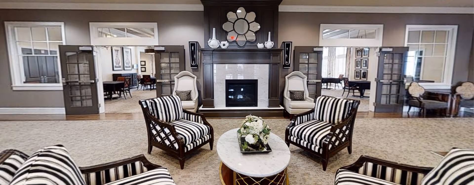 A cozy living room area with a central fireplace surrounded by two white armchairs and four striped armchairs arranged around a white marble coffee table with a floral centerpiece. The room has beige carpeting, gray walls, and glass double doors leading to adjacent rooms with dining tables and chairs.
