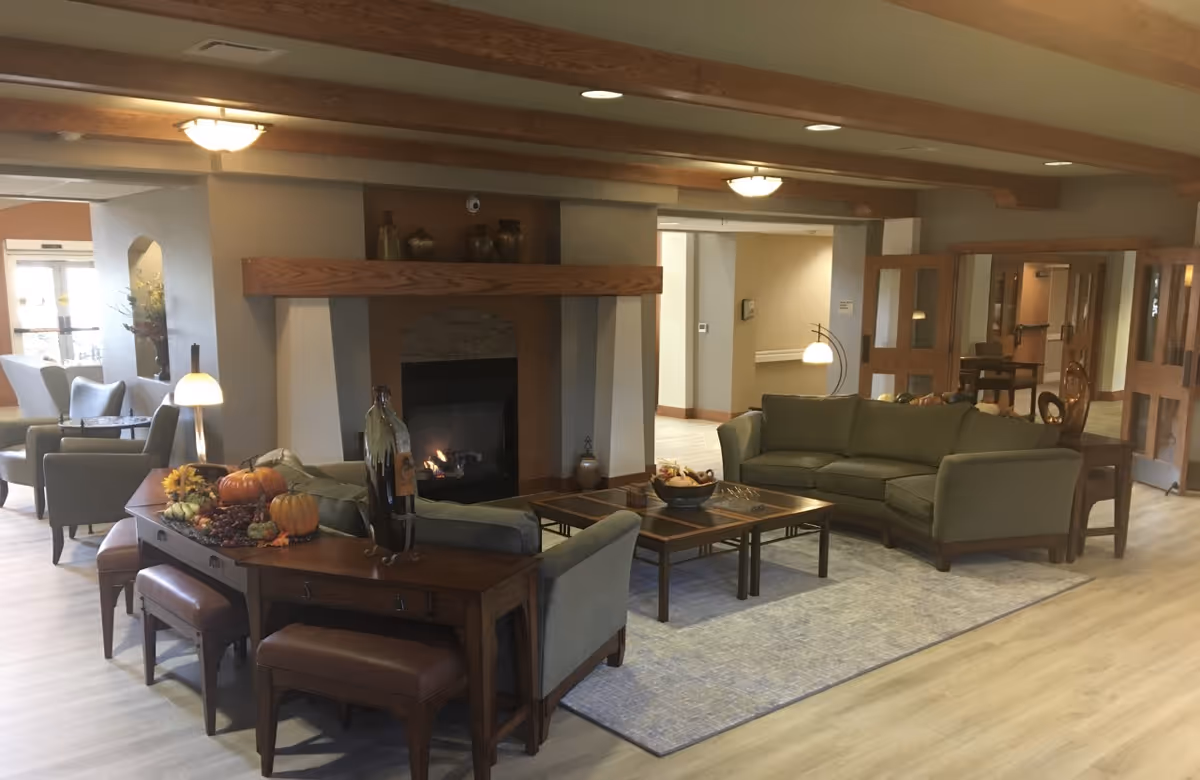 A cozy senior living facility common area with a fireplace, green sofas, wooden coffee tables, and decorative pumpkins on a console table. The room has wooden ceiling beams, light-colored flooring, and soft lighting, creating a warm and inviting atmosphere.
