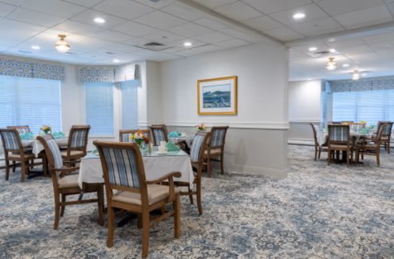 A dining room with several wooden tables and chairs arranged neatly. Each table is set with white tablecloths, green napkins, and small floral centerpieces. The room has large windows with blinds and patterned valances, a patterned carpet, and a framed landscape painting on the wall.
