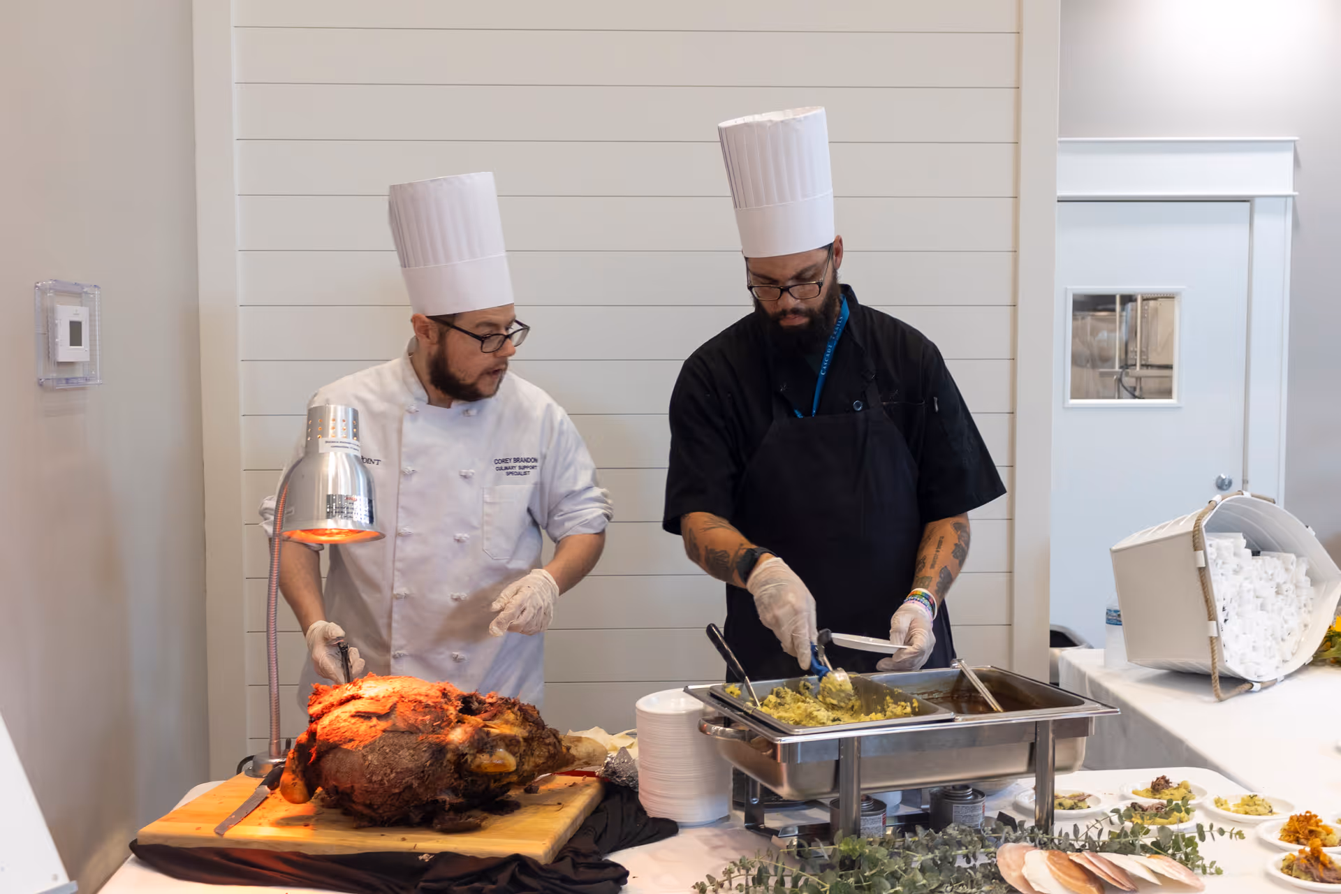Two chefs wearing white chef hats and gloves preparing food at a buffet table. One chef is carving a large roasted meat on a wooden cutting board under a heat lamp, while the other is serving food from a chafing dish. Plates and food items are arranged on the table in front of them.
