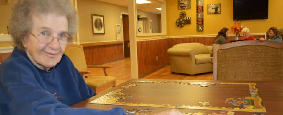 An elderly woman with glasses and curly gray hair is sitting at a table working on a jigsaw puzzle. In the background, three other elderly women are seated on couches and chairs in a cozy room with yellow walls and wooden paneling. The room has framed pictures on the walls and a television mounted above the couch.