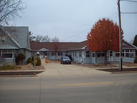 Exterior view of a single-story residential building with gray siding and a brown roof, featuring a driveway with a parked car and a tree with red autumn leaves near the sidewalk.