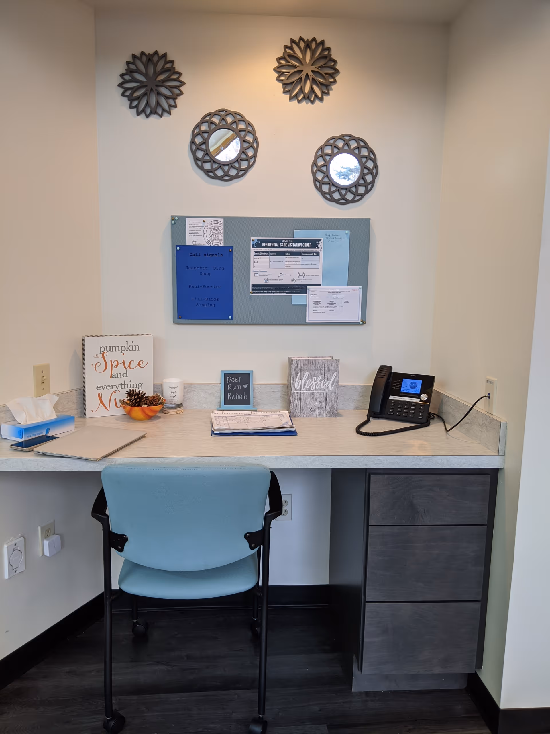 A small office or reception desk area with a light blue chair on wheels. The desk has a phone, some paperwork, a tissue box, a small bowl with pine cones, and decorative signs including one that says 'pumpkin spice and everything nice', another that says 'Deer Run Rehab', and one that says 'blessed'. Above the desk is a bulletin board with papers pinned to it and four decorative round mirrors with intricate frames on the wall.