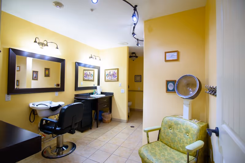 Interior view of a salon area in a senior living facility with yellow walls, a green vintage salon chair with a hair dryer hood, a black salon chair in front of a white sink, two large mirrors on the wall, framed artwork, and tiled floor.