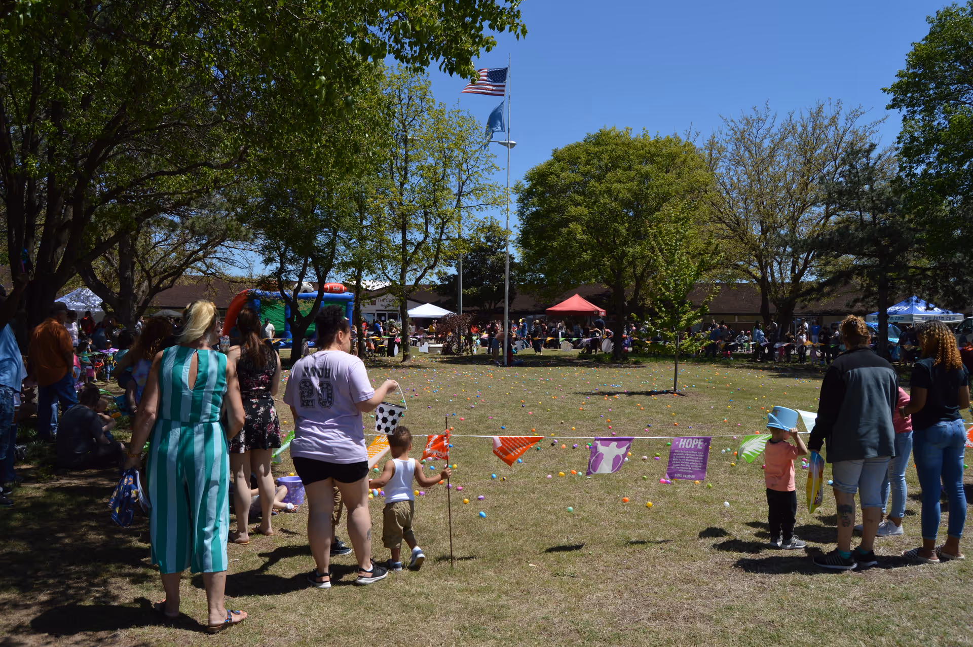 A group of people, including adults and children, gathered outdoors on a sunny day in a grassy area with trees and flags. Colorful plastic eggs are scattered on the ground, suggesting an Easter egg hunt event. Several tents and a bounce house are visible in the background with more people participating in the event.