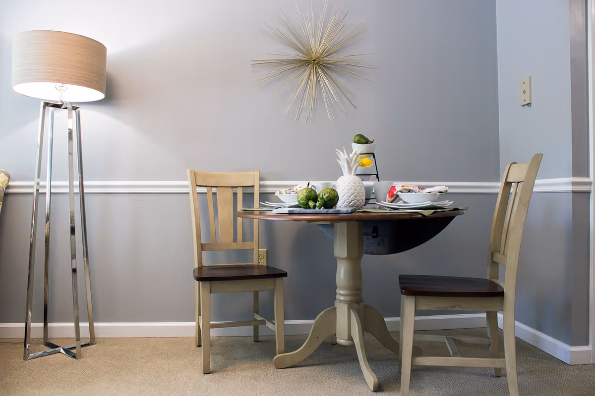 A small dining area with a round wooden table set for two, featuring plates, bowls, and decorative fruit. Two wooden chairs with dark seats are positioned on either side of the table. A modern floor lamp with a beige shade stands to the left, and a gold starburst wall decoration hangs on the light gray wall behind the table.