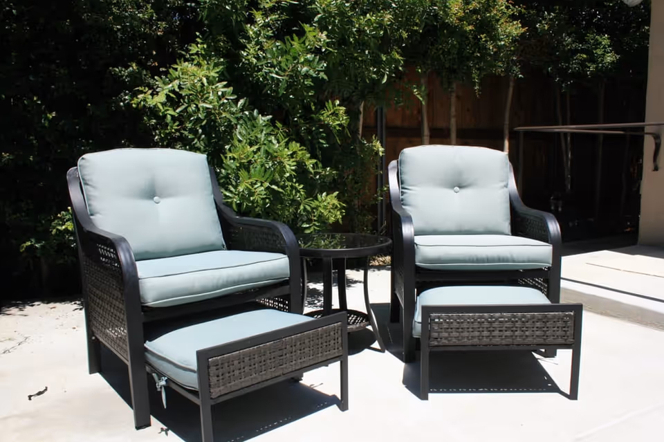 Two outdoor cushioned chairs with matching footrests and a small round glass-top table between them, set on a concrete patio with green bushes and a wooden fence in the background.