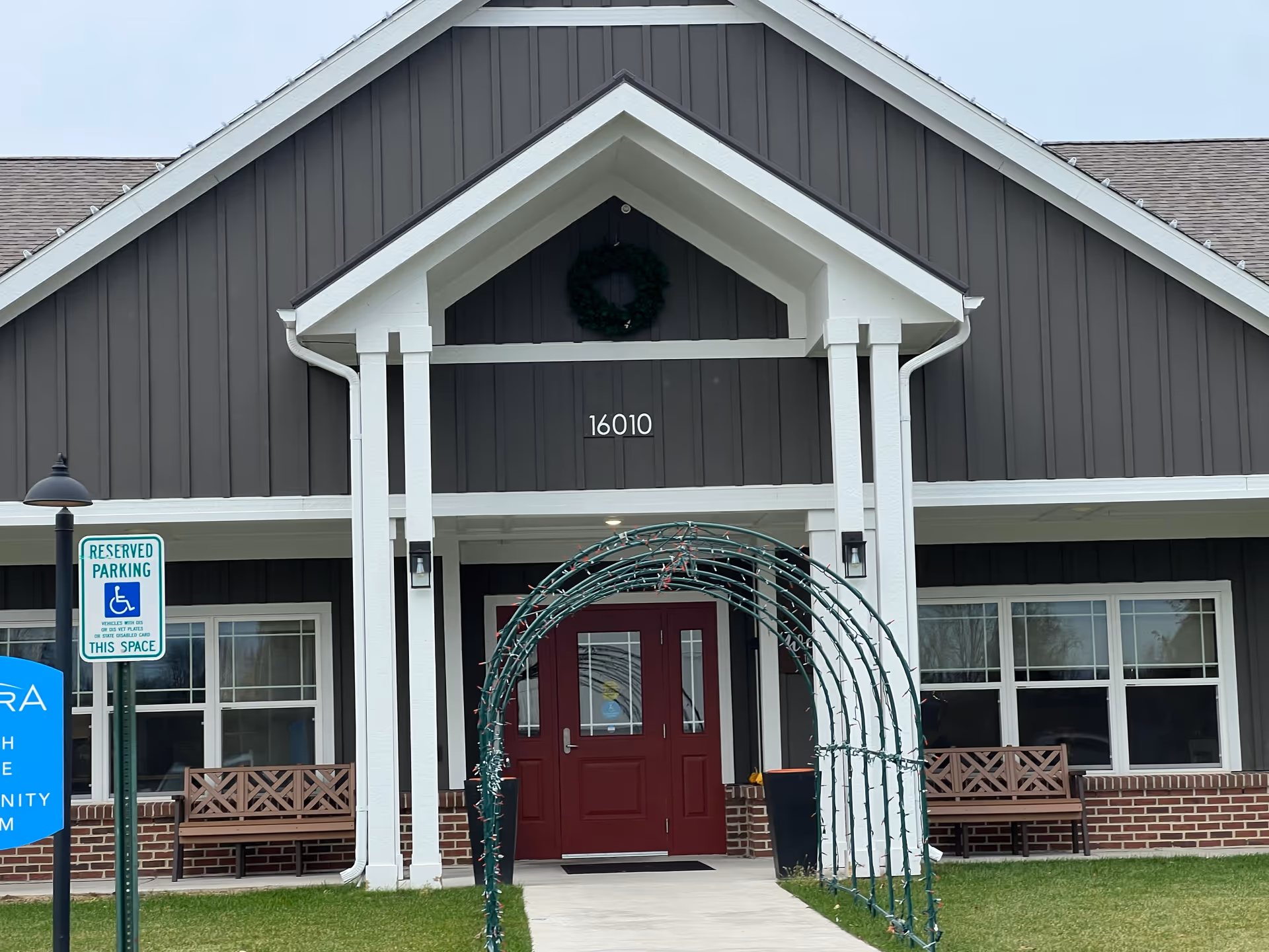 Front entrance of a building with dark gray siding and white trim, featuring a red double door under a covered porch with white pillars. A green archway with string lights leads to the door. There are benches on either side of the entrance and a reserved parking sign for handicapped parking is visible on the left. The building number 16010 is displayed above the door.