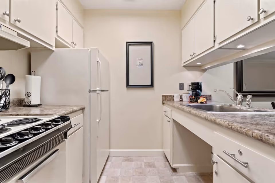 A compact kitchen with white cabinets and beige countertops. On the left side, there is a white refrigerator and an electric stove with black knobs. On the right side, there is a double sink with a chrome faucet, a coffee maker, and some mugs on the countertop. A framed picture hangs on the wall at the end of the kitchen. The floor has light-colored tiles.