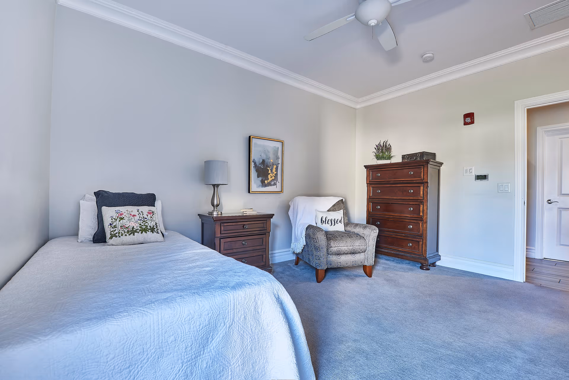 A neatly arranged bedroom with a single bed covered in a white quilt and floral pillow, a wooden nightstand with a lamp and books, a patterned armchair with a white throw and a pillow that says 'blessed', a tall wooden dresser with decorative items on top, light gray walls, and a ceiling fan.