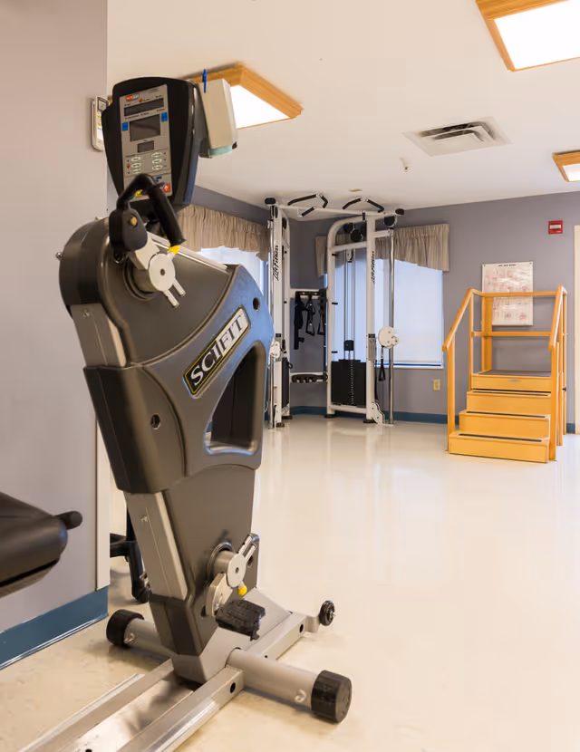 A rehabilitation gym room with exercise machines, resistance equipment, and a set of wooden therapy stairs.