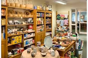 Interior view of a small gift shop or store area within a senior living facility, featuring shelves stocked with various items such as baskets, packaged goods, and pottery. A table in the foreground displays ceramic mugs and bowls, while other tables hold additional merchandise. The background shows a doorway leading to another room.
