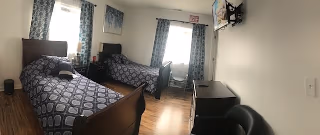 A bright assisted living bedroom with two single beds featuring matching blue patterned bedspreads. The room has wooden flooring, two windows with blue patterned curtains, a small nightstand with a lamp between the beds, a wall-mounted TV, a dresser, and a black chair. An exit sign is visible above one window.