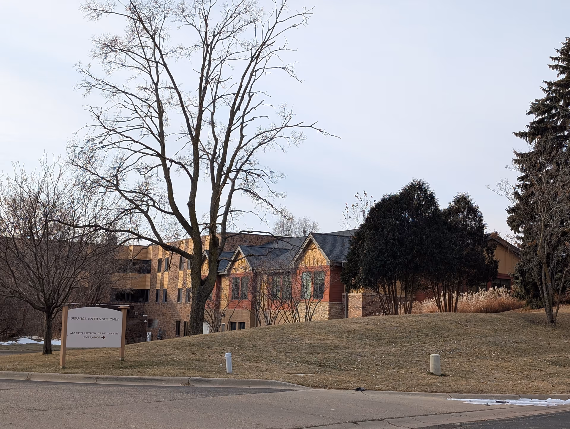 Exterior view of a multi-story brick and wood building partially obscured by leafless trees and bushes, with a sign indicating service entrance and Martin Luther Care Center entrance.