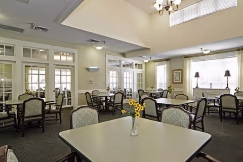 A bright and spacious dining room with multiple square tables and cushioned chairs arranged neatly. The room features large windows with white blinds, allowing natural light to fill the space. There are decorative elements such as a vase with sunflowers on the nearest table, framed artwork on the walls, and two table lamps on a sideboard under the windows. The ceiling is high with a chandelier and recessed lighting, and the walls are painted in a light neutral color.